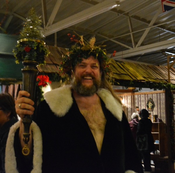 Spirit of Christmas Present costumed characeter with green wreath of candles on his head and fur-trimmed robe holds an evergren tree aloft against the background of the Dickens Fair