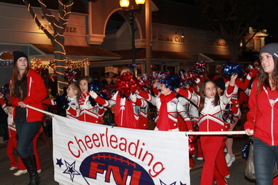 Cheerleaders in red and white outfits wave as they march behind a "Cheerleading FNI" banner.