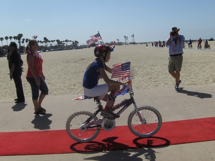 Young boy rides his bike with American flags fluttering from the handlebars down the red carpet to the beach bike path.