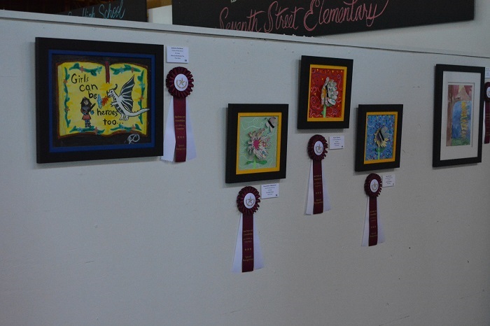 Student art pieces, professionally framed, hand on a white wall with blue ribbons next to them.