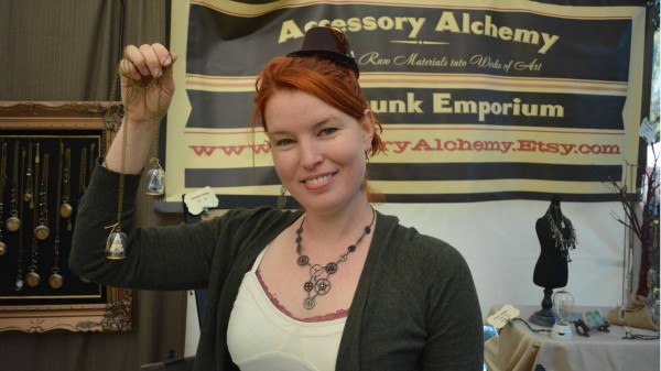 Accessory Alchemy proprietor Ali Krajewski displays her miniature Christmas-tree pendant in front of a banner that says "Accessory Alchemy", at her Jackalope Pasadena booth