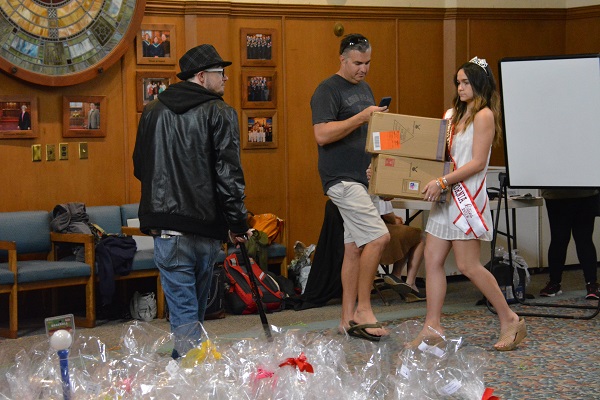 Beauty queen in sash and tiara carries in two boxes of supplies as man photographs her with his phone