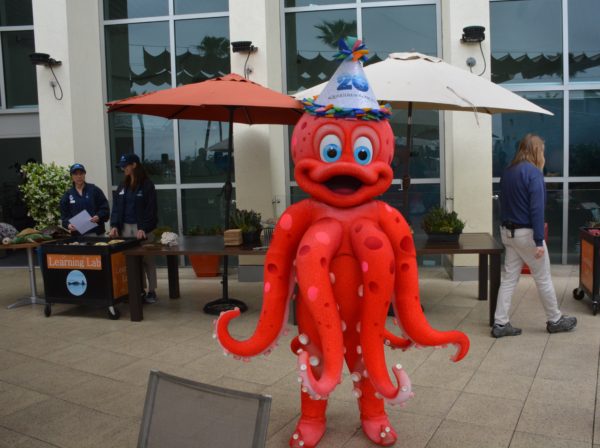 Gigi the red octopus mascot wears party hat as she stnds on Aquarium of the Pacific terrace