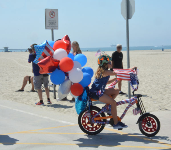 Girl pedals bike decorated with red-white-and-blue balloons and streamers near the beach