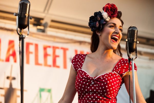 Female vocalist in a 1940-s style red dress with white polka dots sings at a mic in front of a Fleet Week sign.