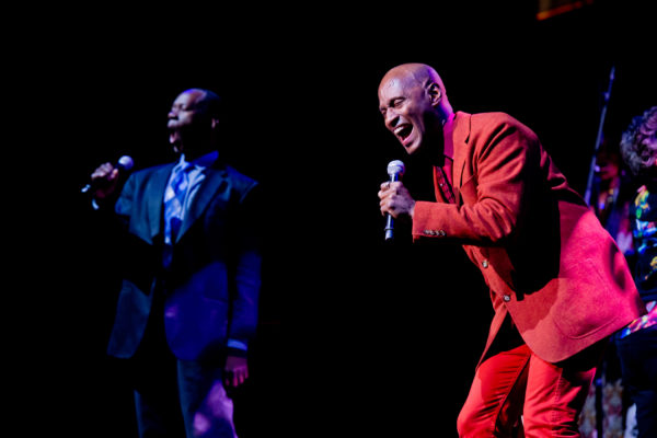 Two male singers onstage, one in a red suit and one in blue, smile as they sing into their mics