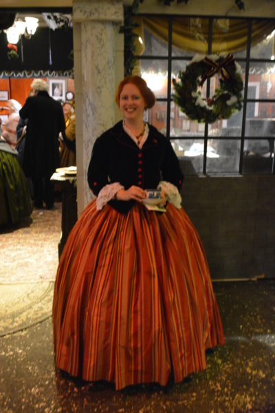 Redhaired woman in a long orange skirt smiles a welcome outside a house with a green wreath in the window