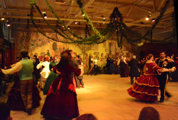 Fezziwigs revelers dance on a wooden parquet floor festooned with greenery