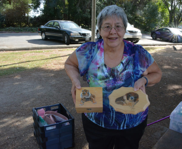 Artist Liz Stark displays a hexagonal plaque with an otter and a square box on which she's carved and painted a tiger, against the background of El Dorado Park