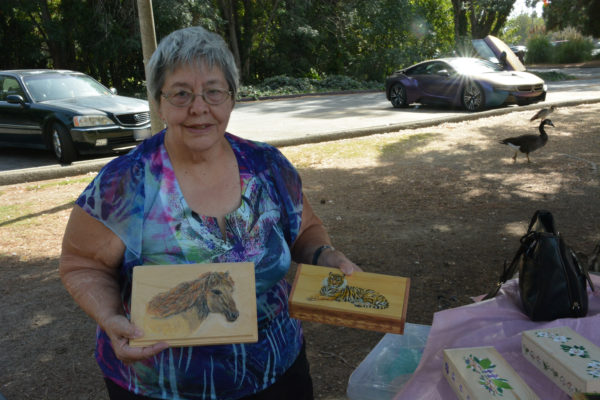 Artist Liz Stark holds up two of her carved and painted boxes against the background of El Dorado Park