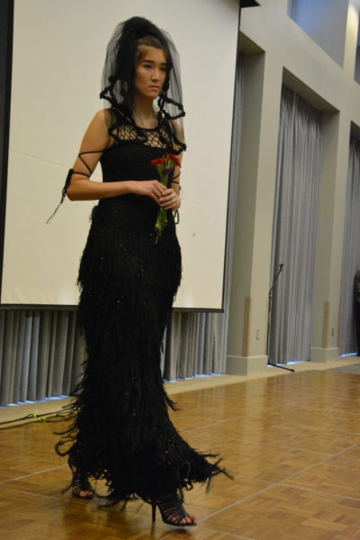 Model exhibits black crocheted wedding dress with fiber fringe on the skirt, veil and stiletto heels at fashion show