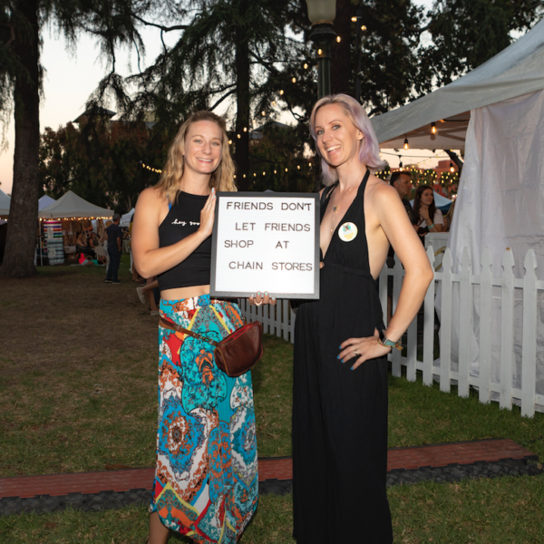 Sara Diederich and Melissa Shipley in long dresses with sign that says, "Friends don't let friends shop at chain stores", near Jackalope Pasadena tents at sunset