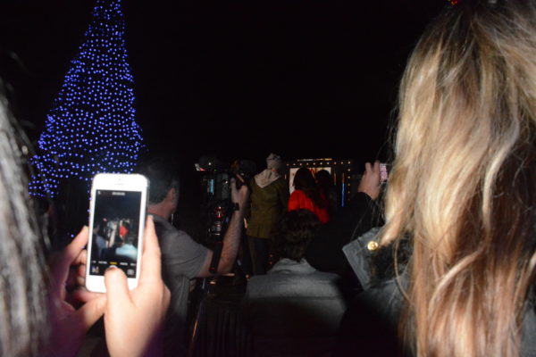four hosts reach to turn on giant Christmas tree's lights as audience members photograph them on their cell phones