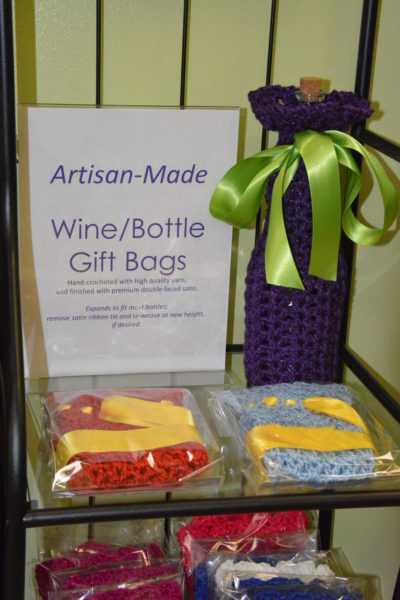 Purple crocheted cover with green ribbon on a wine bottle with two others nearby on shelf and "Artisan-Inspired Wine Bottle Gift Bag" sign