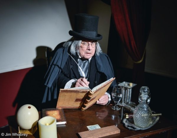 Phil Soinsky, in Victorian top hat, sits behind a table with a ledger and quill pen as "Scrooge"