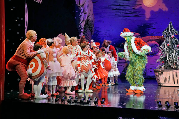 Edward Watts, as "The Grinch", onstage in Santa hat and red jacket with vairous "Whos" in fluffy Christmas dresses and suits