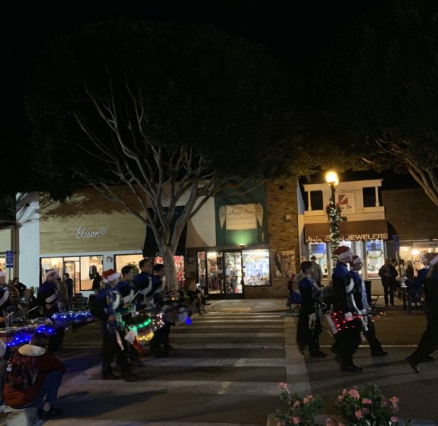 High school students in Santa hats walk by on Seal Beach lighted street at night with instruments, including a drum decorated with green Christmas lights