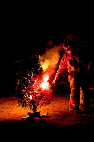 Firefighter stands in the red glow of burning Christmas tree on wooden stand as bystanders look on during Julefest "Safety Burn" event
