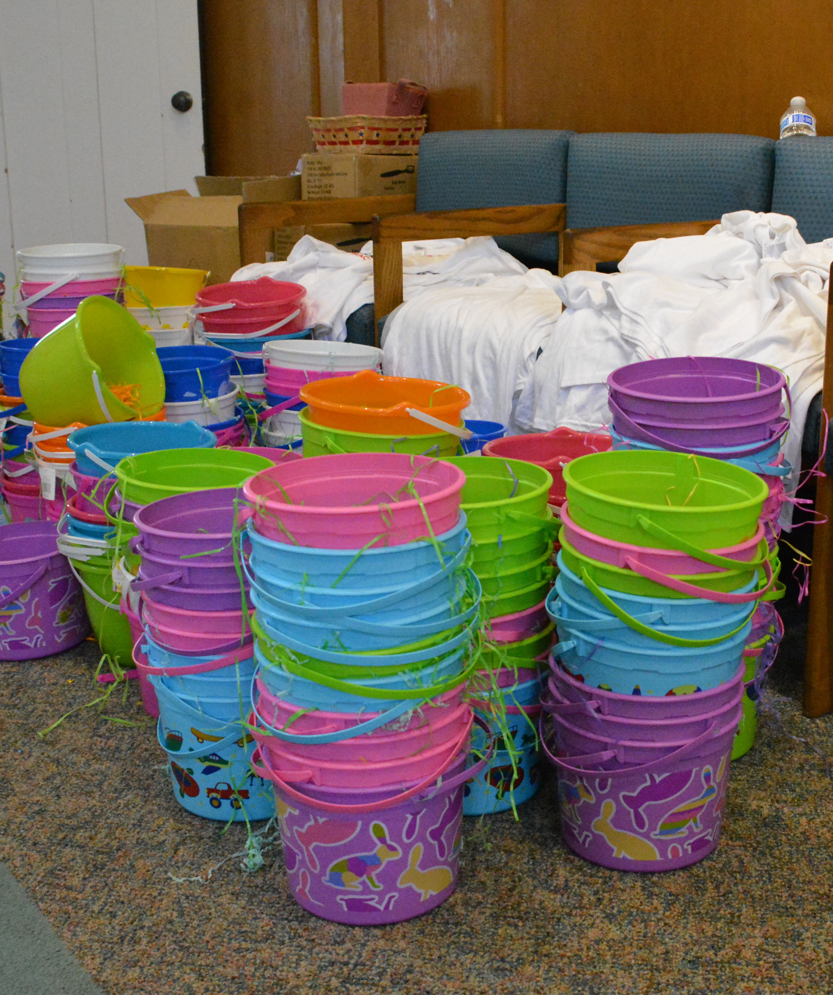 Multicolored plastic sand pails with Easter grass in them sit on carpeted floor waiting to be filled
