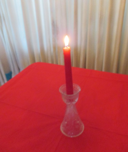 Red lighted taper candle in cut-glass candleholder on red tablecloth near a white curtain