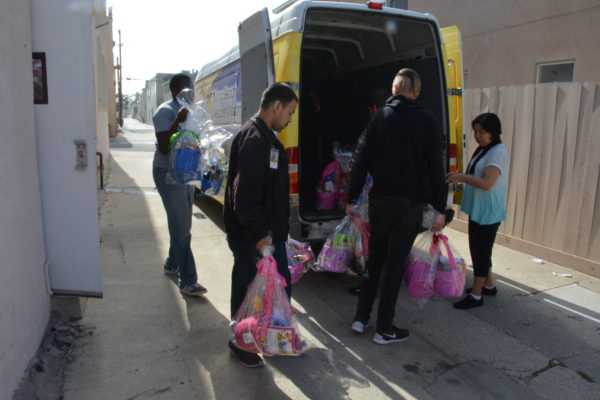 Three volunteers carry completed cellophane-wrapped Easter baskets to a waiting Long Beach Rescue Mission van in an alleyway as a woman supervises
