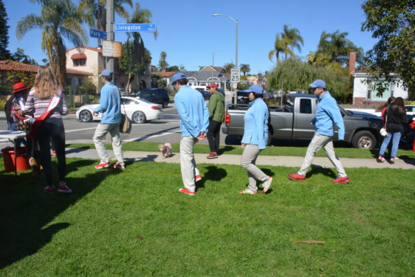 Four people in blue Honda shirts and caps walk across the grass to the pavement, wearing red shoes