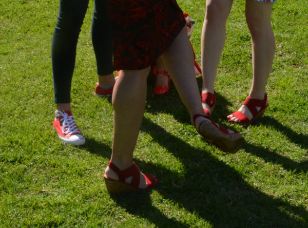 Woman kicks her foot in red platform matching another pair nearby as red sneakered feet stand in background on grass