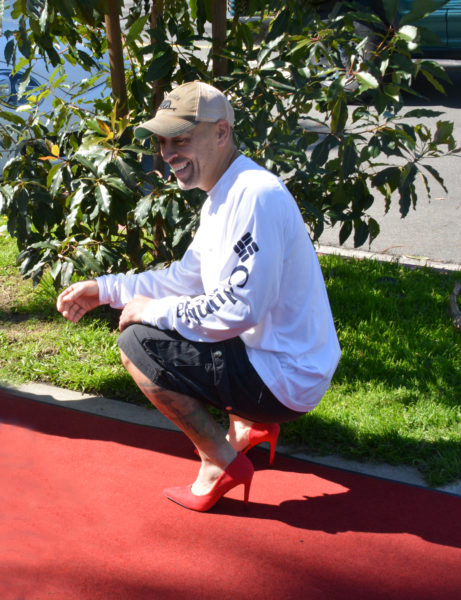 Man in red heels crouches on red carpet and laughs