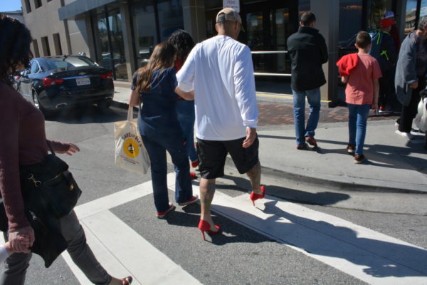 man in red high heels crosses street supported by female companion