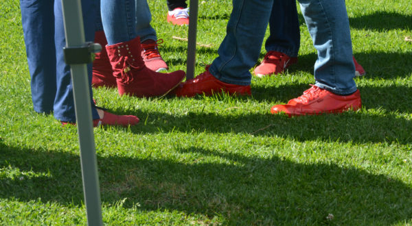Red boots "play footsie" with a foot in a red sneaker with other red shoes on bystanders on the grass 