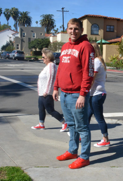 Man in red"Washington State" sweatshirt smiles as he stands on street corner in spraypainted red shoes as two women in red sneakers walk by