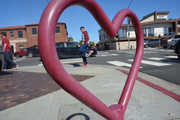 teenage boy in red shoes lifts foot in a playful kick framed by red iron heart scuplpture on street