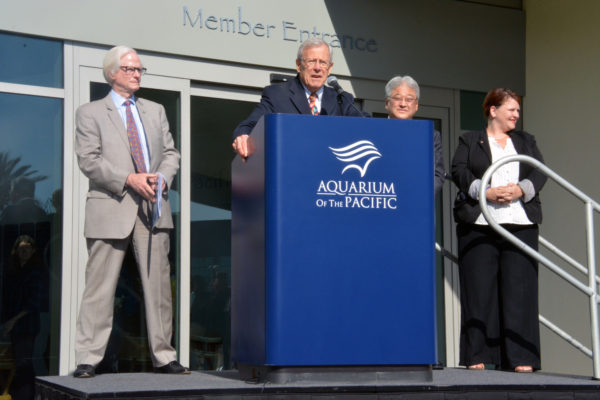 Dr. Jerry Schubel speaks from a blue podium with "Aquarium of the Pacific" on it in white letters, flanked by Doug Otto, Steve Morikawa and Jeannine Pearce