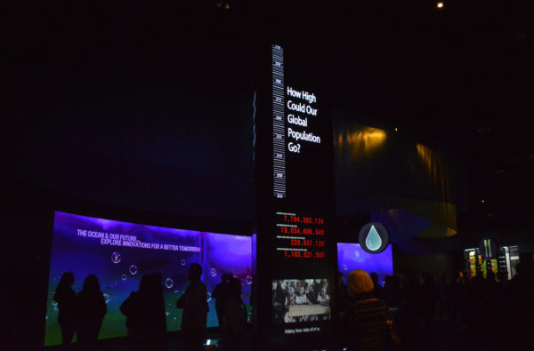 visitors in silhouette next to interactive screen beyond "thermometer" display with the words, "How high could our global population go?"