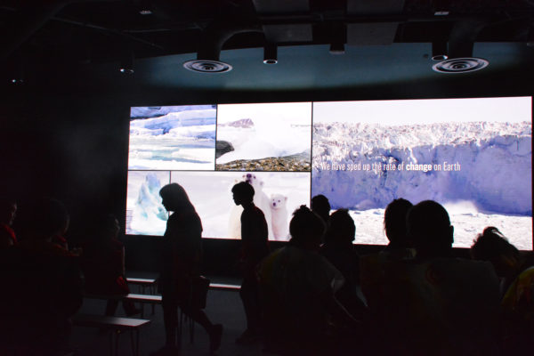 Students silhouetted against a screen with graphics of melting polar ice caps and the words, "We have accelerated the rate of change on earth".