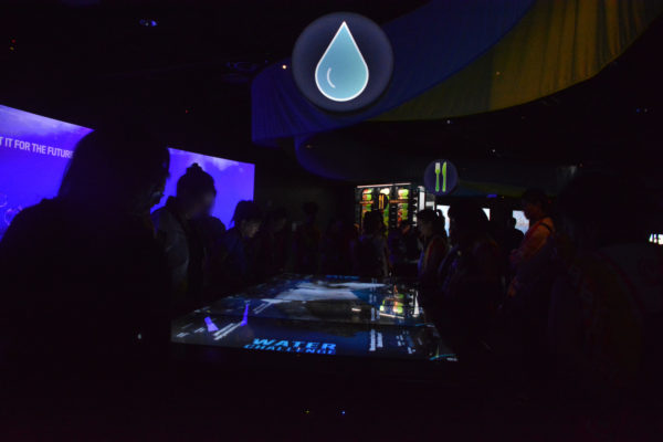 High school students gather around interactive lighted display with "WATER CHALLENGE" on it and a blue-lit water drop overhead