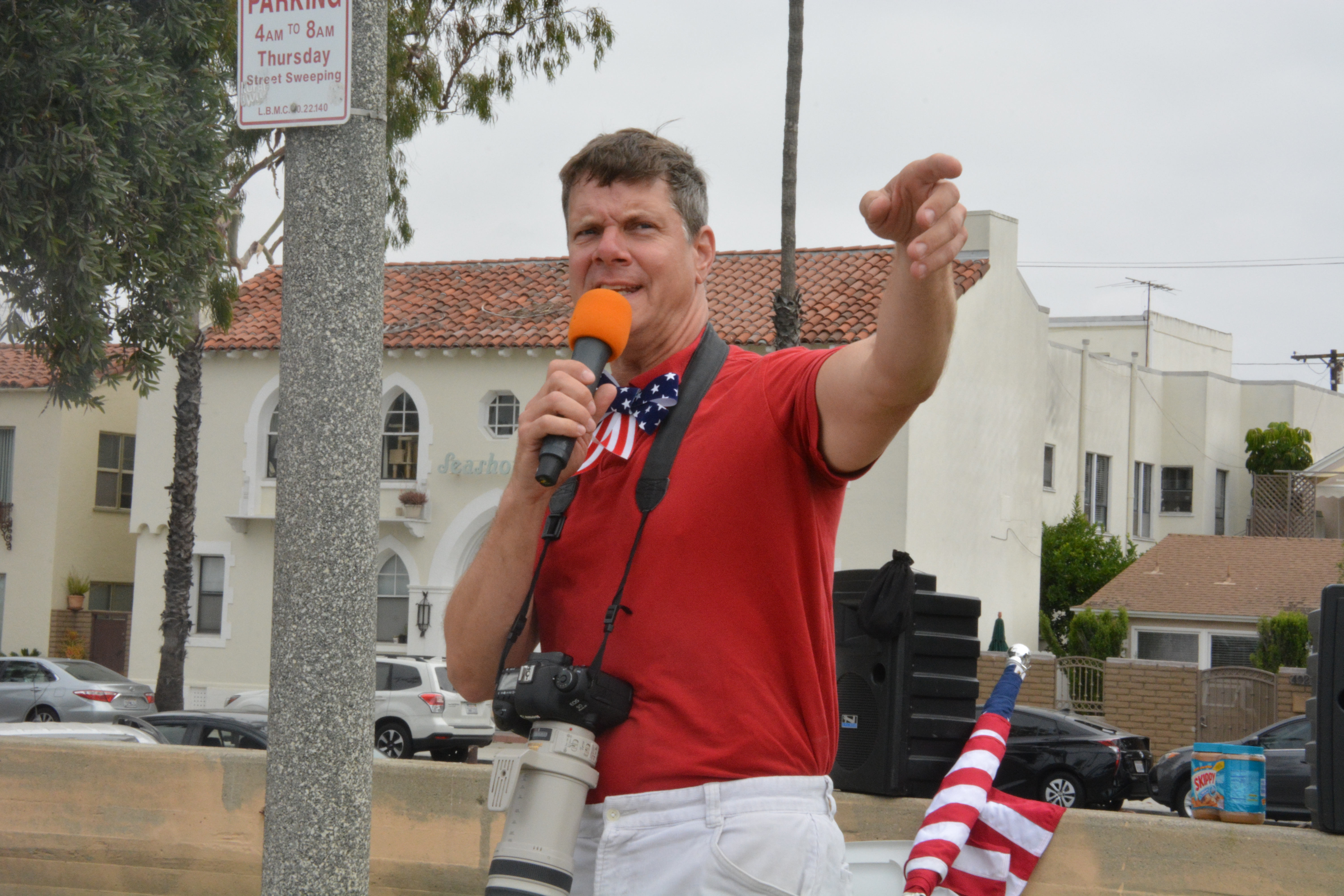 Community activist Justin Rudd wears a camera around his neck and a red-white-and-blue bow tie over his red polo shirt as he speaks on mic and points towards the crowd