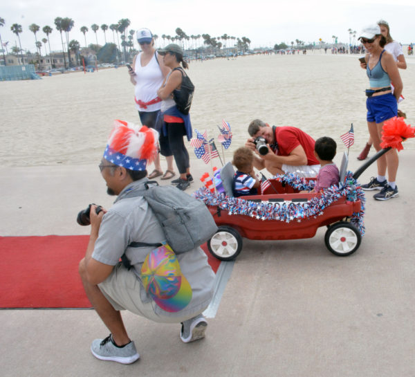Justin angles his camera at two babies in a decorated red wagon while photographer with red-white-and-blue headdress squats near red carpet