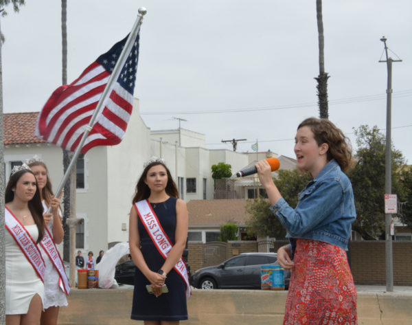 Shannon Wynn sings into a hand-held microphone as three beauty queens stand nearby with the American flag