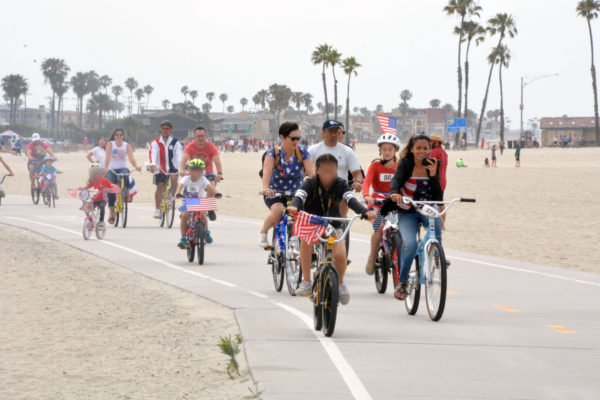 Adults and children ride red-white-and-blue decorated bikes on bike path near the ocean with palm trees in the background