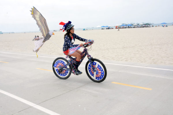 young girl in a blue shirt with white stars rides a bike with red-white-and-blue streamers twined through its spokes and an eagle balloon tied to its seat