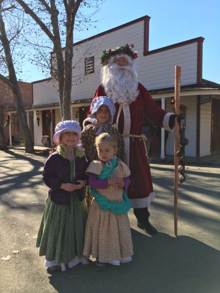 Father Christmas with a wreath on his head, stands near two little girls in period costumes