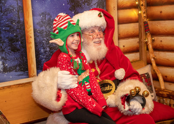 Small girl smiles from "Santa's" lap while wearing an elf-ears cap near log-cabin background