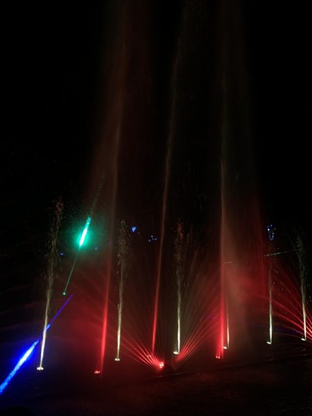Multiple tall red and gold cones of light, highlighted by a green spotlight, rise through jets of water