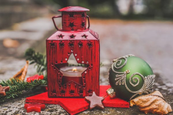 Red metal lantern with star-shaped opening and candle inside on red cloth with a star ornament and green Christmas tree ball nearby