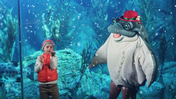 Child looks at a handful of snow next to aquarium tank and stingray character with red hat