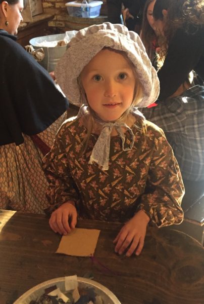 Small girl in period costume and bonnet works with craft supplies 