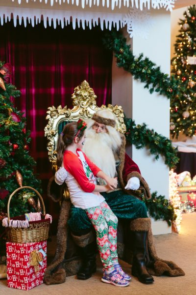 Santa talks to a girl of about nine as he sits on a throne near a Christmas tree and a pillar wrapped in greenery