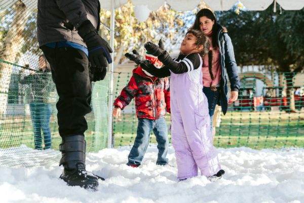 Small girl tosses a handful of snow into the air as small boy walks across snow and their parents stand nearby in snow play area