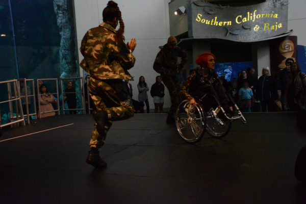 Auti Angel does a dance maneuver in curstom wheelchair with silver dollar signs on its spokes as dancer Ossi leaps nearby